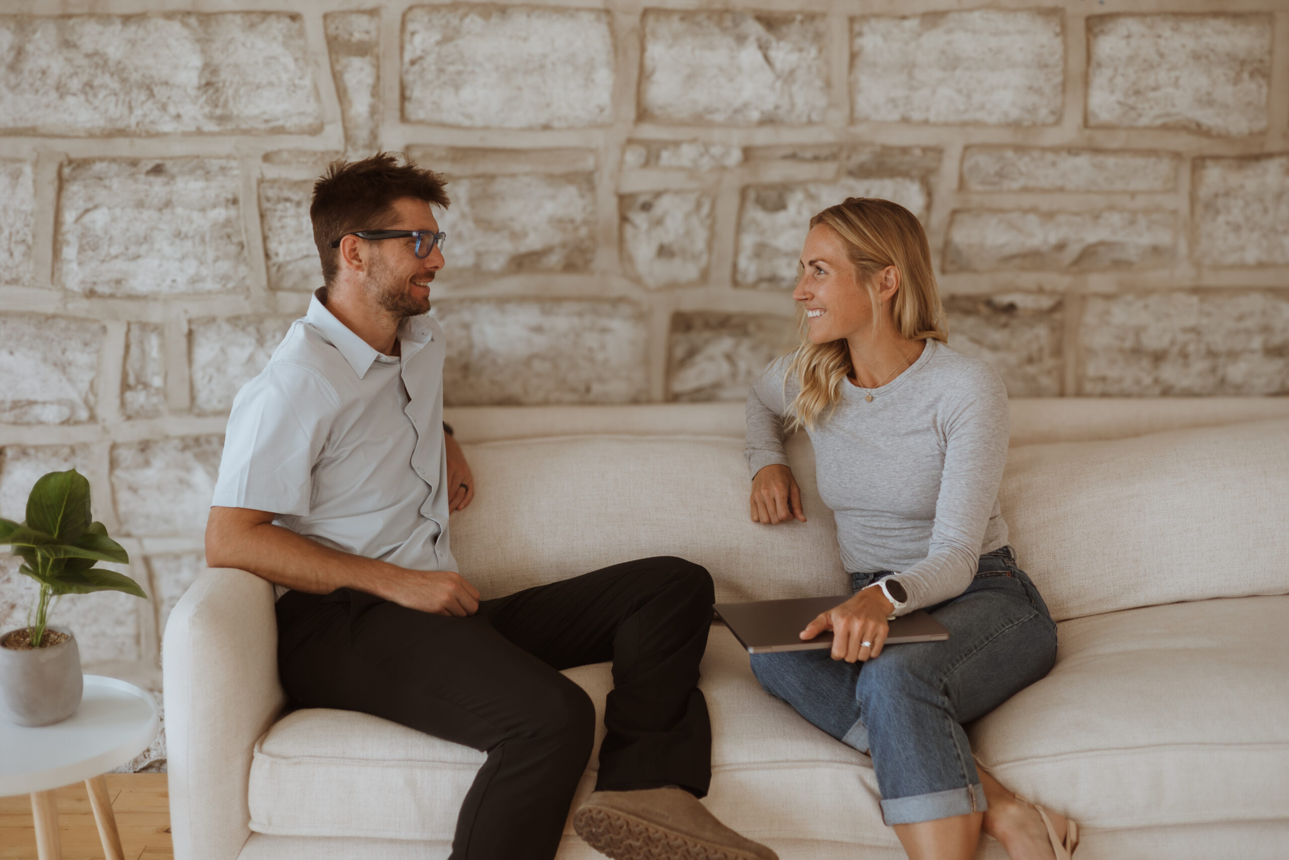 Brant Stachel and Elaina Raponi sitting on a couch, smiling at each other, with a stone wall in the background.